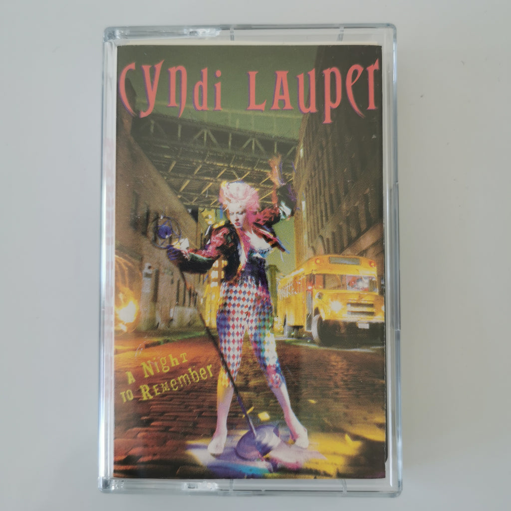 A clear plastic cassette case containing Cyndi Lauper's album 'A Night to Remember', featuring a colorful cover illustration of a woman in checkered clothing standing in a city street with a yellow bus in the background.