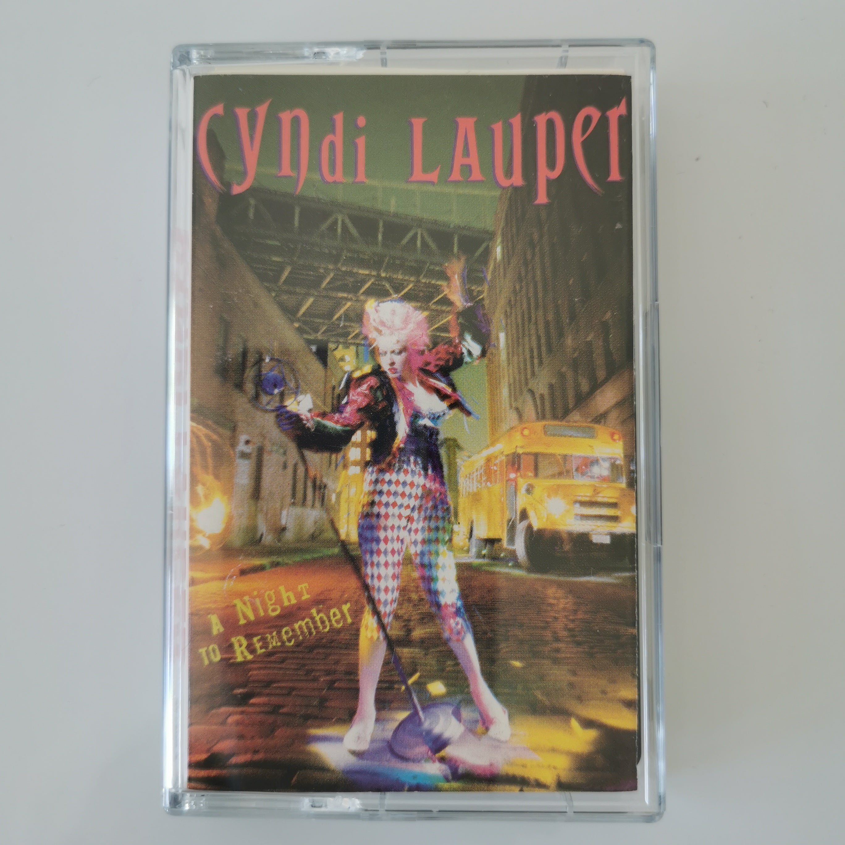A clear plastic cassette case containing Cyndi Lauper's album 'A Night to Remember', featuring a colorful cover illustration of a woman in checkered clothing standing in a city street with a yellow bus in the background.