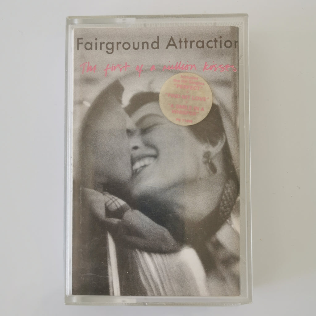 A cassette tape of Fairground Attraction's 'The first of a million kisses' with a black and white photo of a smiling woman on the cover, featuring a pink handwritten title and a gold sticker.