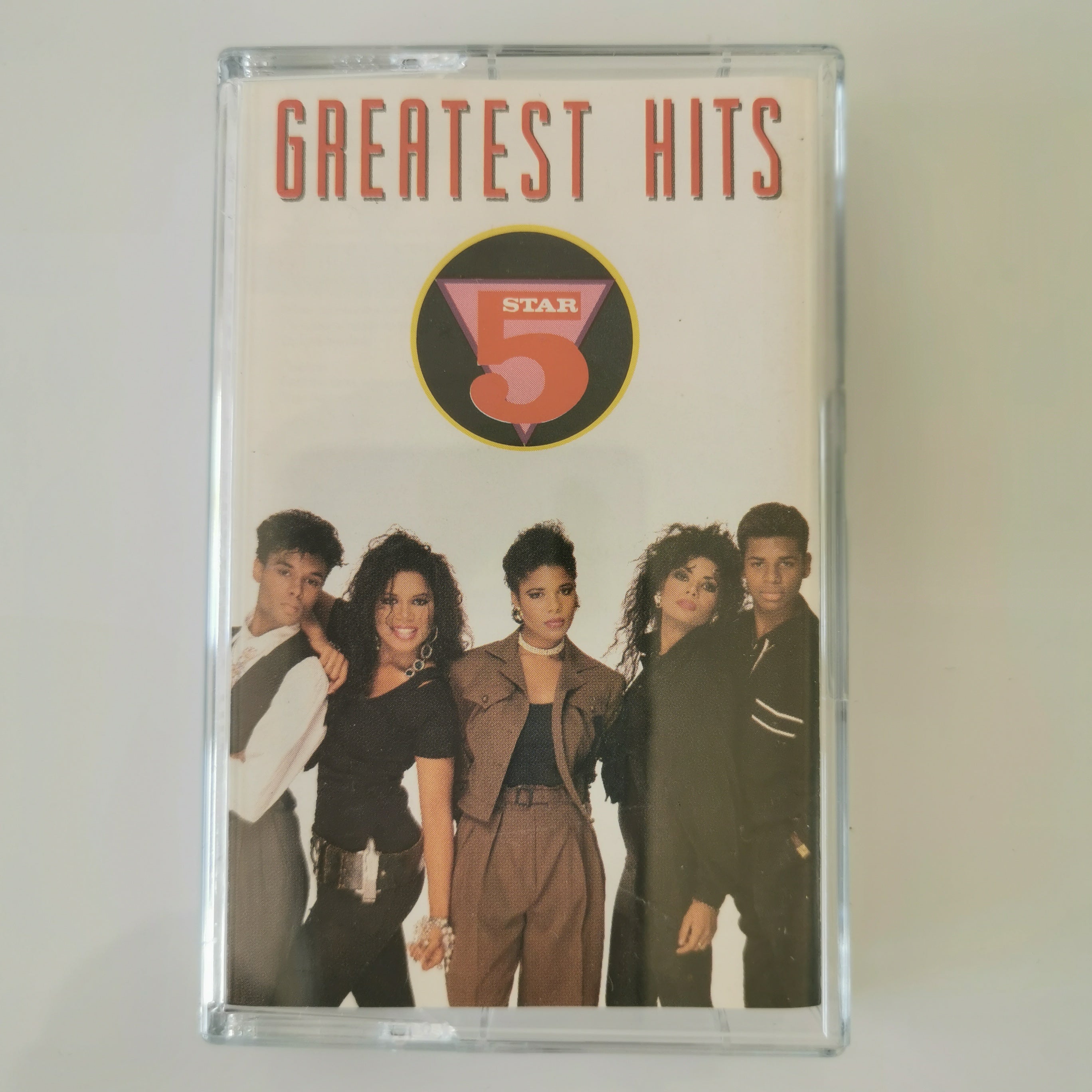 A clear plastic cassette case containing a music compilation album titled 'GREATEST HITS' with a Star 5 logo and a group photo of five singers on the cover, set against a plain light background.