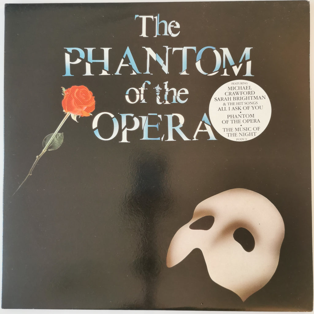 The Phantom of the Opera vinyl record cover, featuring a black background with the title in stylized blue and white lettering, a red rose on the left, and a cream-colored mask in the lower right corner.
