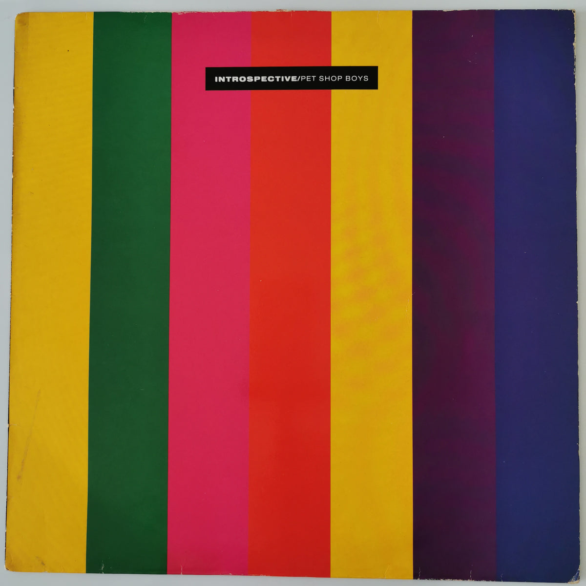 A square album or book cover featuring bold vertical stripes in yellow, green, pink, red, orange, purple, and blue, with a small black label near the top reading 'INTROSPECTIVE/FT SHOP BOYS'.