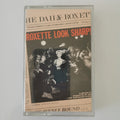 A clear plastic cassette case containing a music compilation titled 'The Daily Roxette' with cover art featuring the text 'Roxette Look Sharp!' and a black-and-white crowd scene, photographed against a light neutral background.