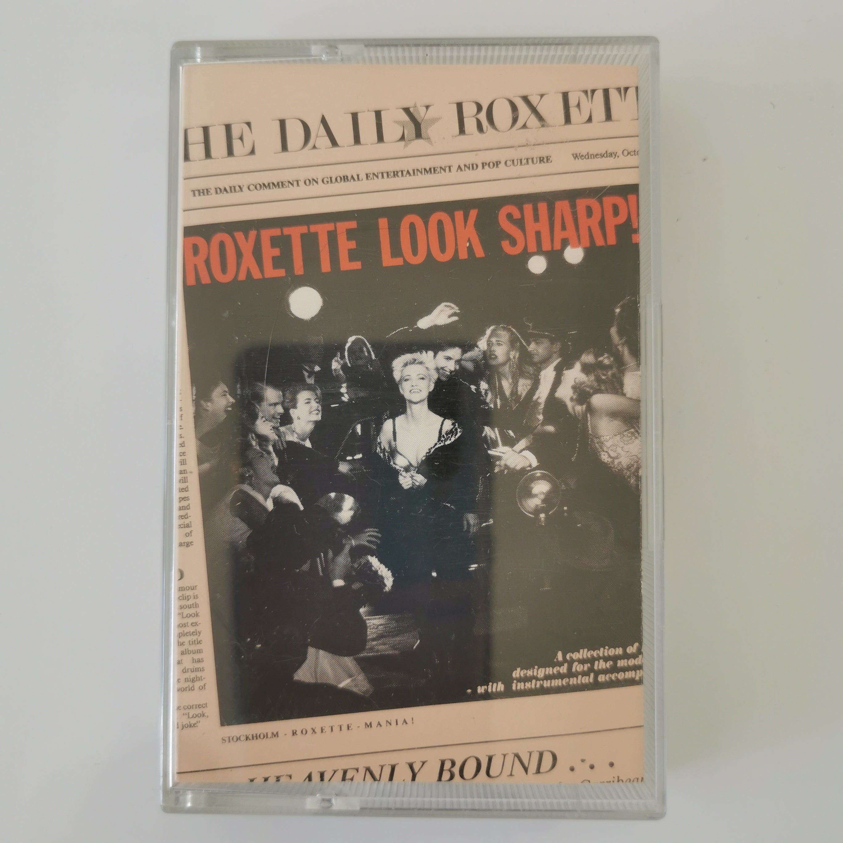 A clear plastic cassette case containing a music compilation titled 'The Daily Roxette' with cover art featuring the text 'Roxette Look Sharp!' and a black-and-white crowd scene, photographed against a light neutral background.