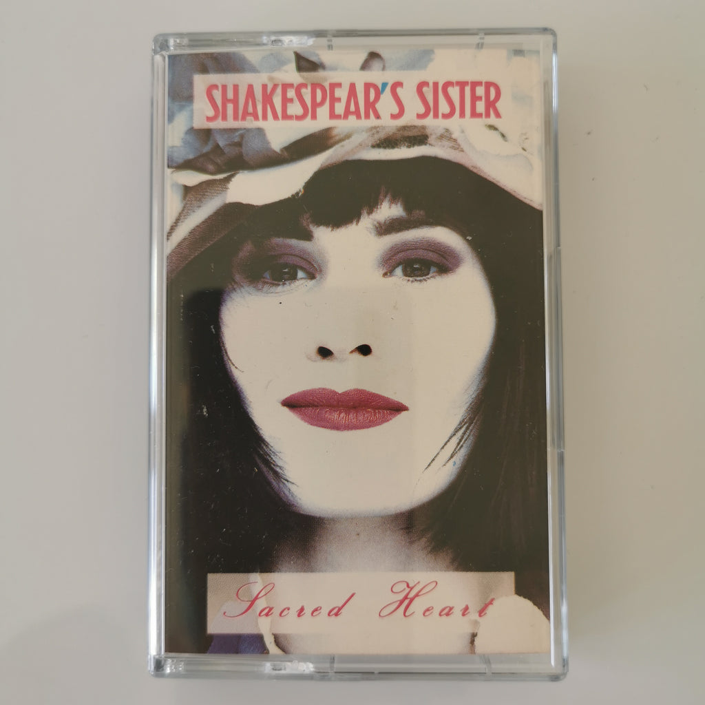 A cassette tape of Shakespeare's Sister's 'Sacred Heart' album, housed in a clear plastic protective case, photographed against a neutral light gray background.