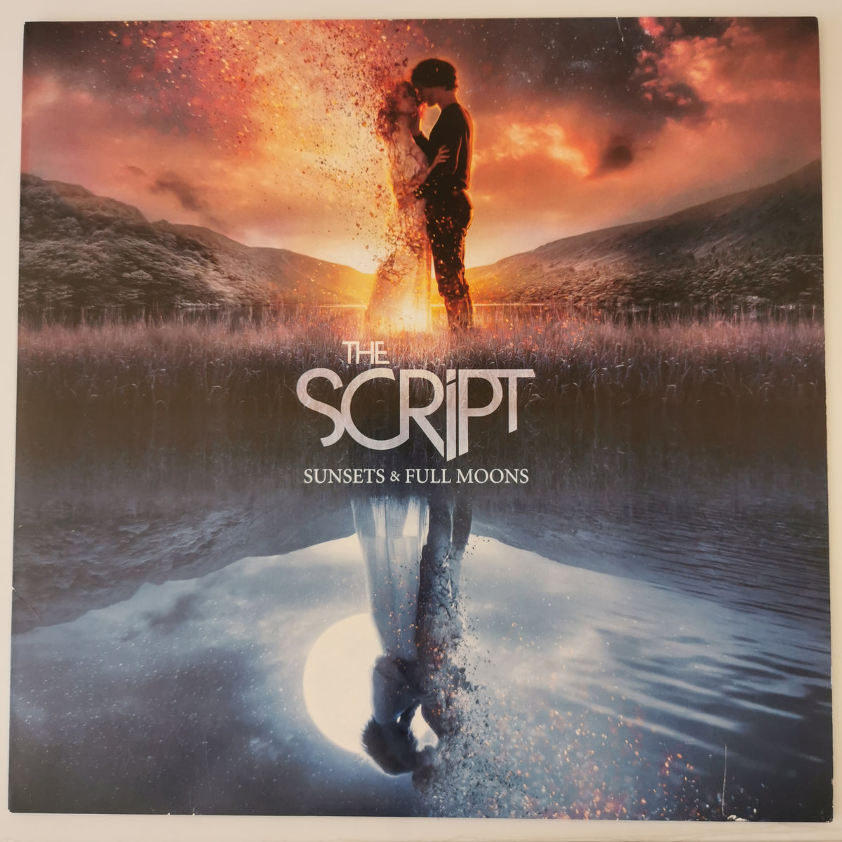 Album cover for 'The Script - Sunsets & Full Moons' featuring a dramatic sunset scene with a couple embracing, their figures dissolving into sparkles, and a reflection in water below.