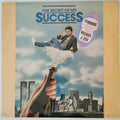 The Secret of My Success motion picture soundtrack vinyl record cover, featuring a illustrated scene of Michael J. Fox floating above a city skyline while a hand holds a champagne bottle.