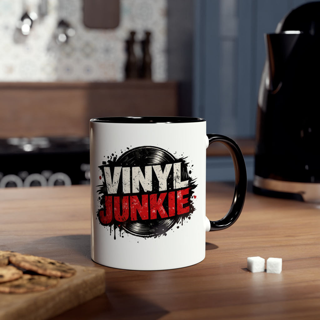 A white ceramic mug with a black handle and interior, featuring a graphic print that reads 'VINYL JUNKIE' in white and red over a black vinyl record design. The mug is placed on a wooden table in a kitchen setting with cookies and sugar cubes nearby.