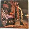 A close-up photograph of a heavily customized motorcycle at night, featuring a purple license plate with 'Ryzen' in white script, a red and black studded leather body cover, and a person's leg in black leather pants and boots resting near the exhaust. The photo has a vintage, grainy aesthetic with warm, orange-toned streetlights.